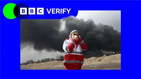 An emergency worker in Tehran shields her face as black smoke rises behind her. She is wearing a red and white jacket and a face mask and standing in a sandy location. The image is imposed over the BBC Verify colours and branding. 