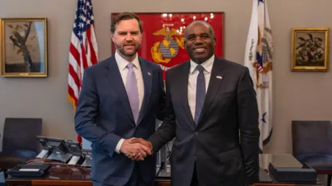 JD Vance and David Lammy shaking hands in an office, in front of two flags – one embroidered with the American stars and strips and another with the bald eagle. 