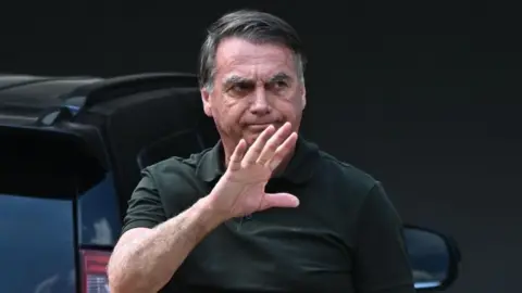EPA A shot of Brazil's former president Jair Bolsonaro waving to the right, dressed in a dark green shirt and hair combed to the right, while walking in front of a car.