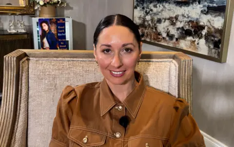 A woman with dark hair tied back is looking into the camera. She has dark eyes and red lipstick. She is wearing a light brown to with white buttons and is sitting on a beige seat.