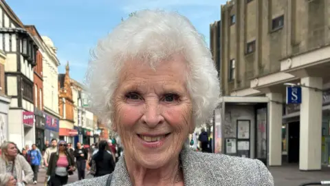 PA Media Margaret Wilson smiles at the camera. She has short grey hair and wears a grey blazer. People can be seen walking behind her through Bedford
