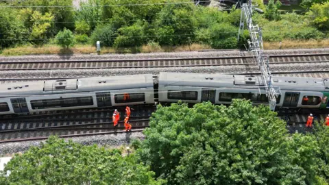 An aerial view of the train showing the first carriage pointing to the right slightly off the main track. There are four sets of rails and a gantry overhead. Several workers in orange high vis clothes mill around. Trees and shrubs are either side of the tracks.