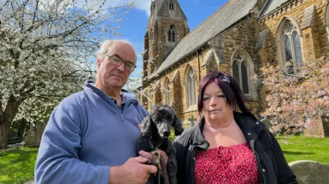 Philip Wallis and Kate Collins are standing in the green grounds of a grand church with yellow and red cobbled brickwork. Philip is holding a black dog and is wearing a blue jumper. Kate on the right is wearing a red dress and black coat.