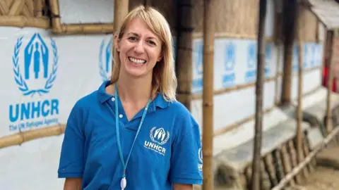 Amber is wearing a blue UNHCR polo shirt stands in front of a row of bamboo-framed shelters covered with white tarpaulin displaying the UNHCR logo and text “The UN Refugee Agency.” The individual has a lanyard with an identification badge and is dressed in green trousers. The setting appears to be an outdoor area with a dirt pathway and trees visible in the background.