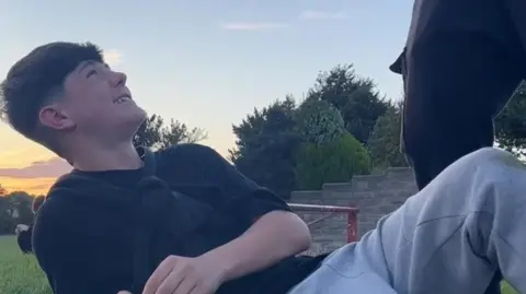 FAMILY PHOTO Osian lays on the grass smiling at his friends at sunset