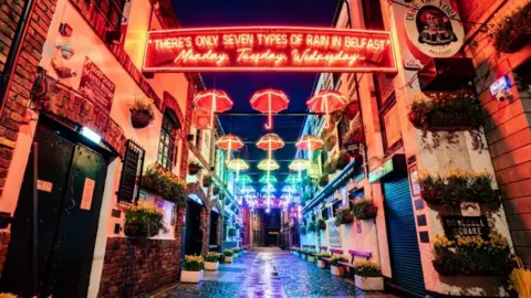 A cobbled street with rows of brightly coloured neon light up umbrellas above head.
