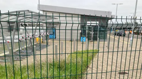 A fenced-off area outside a small modern station building in Winslow, Buckinghamshire, with bike racks on the left and ticket machines near the entrance.