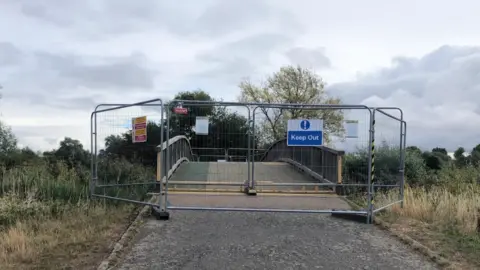 A wooden bridge with a temporary grey wire fence in front of it. There is a tarmac path leading up to the bridge and a blue sign on the metal fence saying "Keep Out".