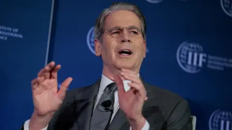 Scott Bessent raises his hands while talking, sitting in front of a dark blue screen.