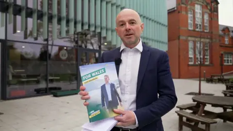 Gareth Lewis in a jacket and white shirt holding up a manifesto with a picture of Rhun ap Iorwerth saying "For Wales"