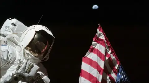 NASA Harrison Schmitt is photographed next to the United States flag on the lunar surface during the Apollo 17 mission. The highest part of the flag appears to point toward our planet Earth in the distant background. Its red and white stripes are also reflected in the visor of Schmitt's helmet.