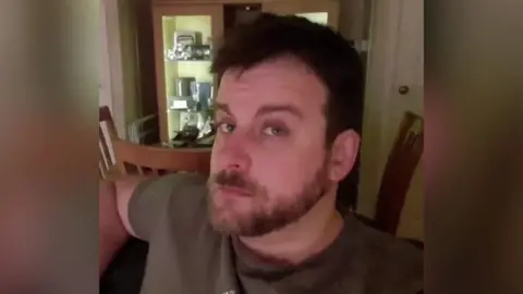 A man with brown hair, a beard and moustache looking at the camera. He is wearing a brown t-shirt. Behind him there are three wooden chairs and a cabinet. 