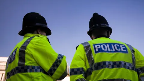 Two police officers wearing black hats and high vis jackets have their arms crossed and their backs towards the camera. 