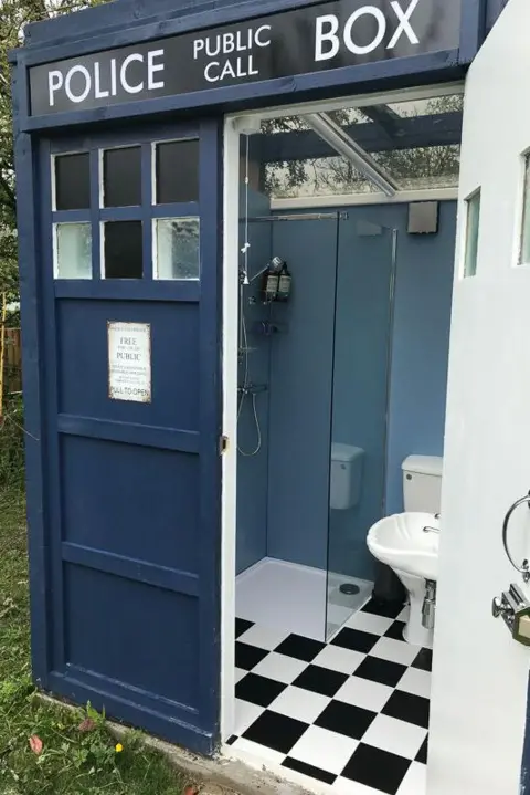 Melin Mabes A replica of Doctor Who's time machine, converted into a bathrom. The outside is blue, beyond the open door we can see a glass shower, white toilet and a basin. The floor is chequered black and white. 