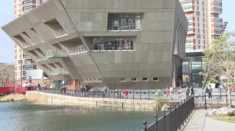 A building with slopey silver-coloured walls overhangs some water with railings around it, people are walking around it.