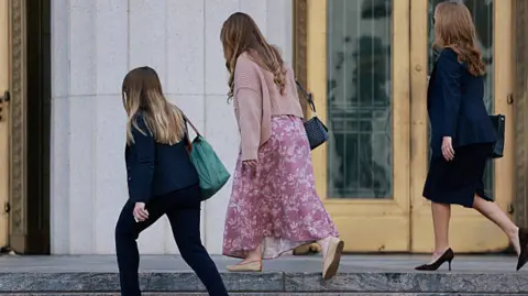 Three women seen from behind are approaching a building. One is shorter and wearing a dark trouser suit, the middle one is wearing a floaty dress and the one on the right is wearing a dark skirt suit. 