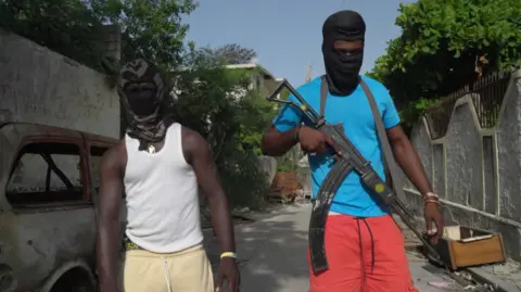 BBC/ Phil Pendlebury Two young men with their heads and faces covered by black hoods and masks, except for their eyes, facing the camera. The one on the right is wearing a blue T-shirt and red shorts, and carrying a large gun. The one on the left is in a white vest and pale yellow shorts.