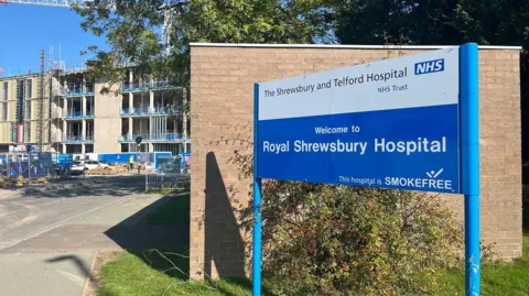 A blue and white sign that says Welcome to Royal Shrewsbury Hospital is the focus of the image. It stands in front of a wall, and behind that in the background, to the left, a four storey building which is still under construction can be seen