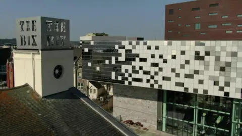 An aerial shot of the Box museum and art gallery in Plymouth taken on a clear sunny day. The words 'The Box' are visible on top of one of the buildings while the main large builder is in the background with big glass windows. 