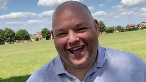 Gary Stanley, standing outside, by a field, smiling. He is wearing a blue top. Buildings and trees are behind him in the field. 