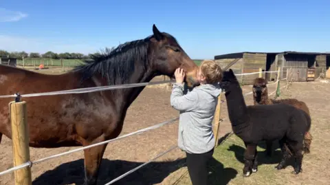 Julia Stewart - a woman with blonde hair and wearing grey hoodie - is stroking and kissing a large dark brown horse. On her right are two dark brown alpacas. There are wooden sheds behind her and blue skies.