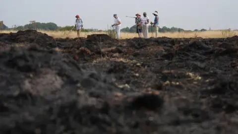 A group of walkers stop to look at the aftermath of a grass fire in Wanstead Flats, north-east London. The fourth heatwave of the summer will continue with temperatures expected to climb to 34C in parts of England. Picture date: Wednesday August 13, 2025. 
