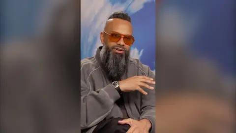 A man with orange shades and a long black beard raises his hand mid speech.