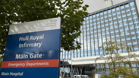 A multi-storey hospital building with blue cladding. A few trees and vehicles are in front of the building.