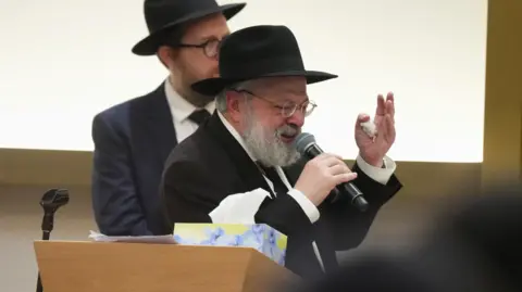 Rabbi Ulman speaks into microphone whilst crying