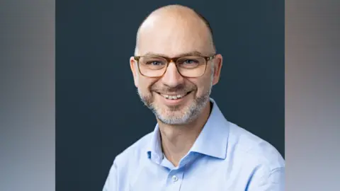 Riverlane Steve Brierley smiling and looking directly towards the camera. He is wearing a light blue open-necked shirt and rectangular framed glasses. He has a beard, which is flecked with grey hair. He is seated against a dark backdrop.