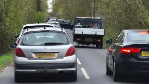 A line of traffic on a narrow, two‑lane road bordered by dense greenery. The road appears to be in a rural or semi‑rural area, with trees and bushes lining both sides and no visible buildings nearby On the left, there is a silver hatchback viewed from behind. The car has a yellow rear license plate. The brake lights are not illuminated.
On the right, there is a dark‑coloured (possibly black) sedan, also viewed from behind. The red rear lights on this vehicle are illuminated, indicating braking or slow movement. Both vehicles are positioned close together, suggesting slow or stopped traffic. Ahead of the foreground cars, there is a small flatbed or light lorry in the same lane as the silver hatchback. The lorry has an open rear bed with metal sides, and objects are visible in the back, though they are not clearly identifiable. Several other vehicles are visible further ahead, forming a queue that extends along the road.
