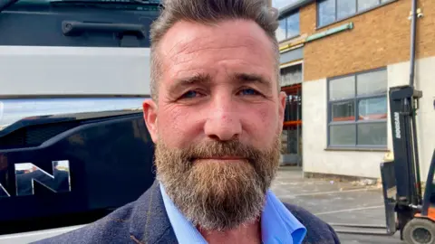 A bearded man stood in front of a lorry in a works yard