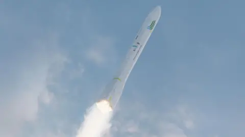 A white rocket flying through the air. Blue sky in the background. There is fire and fuel coming out of the rocket itself. 