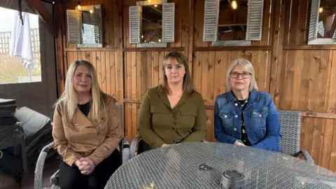 Three women sitting in a row. The first has blonde hair and is wearing a light brown jacket, the second has darker hair with a fringe and is wearing a green shirt and the third has short grey hair that comes to her shoulders and is wearing glasses and a denim jacket. They are sitting in an outdoor seating area with wooden panels behind them.