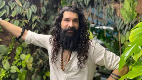 Jonny has long dark curly hair and a beard. He is wearing a white linen shirt and a wooden necklace and is surrounded by all kinds of plants. 