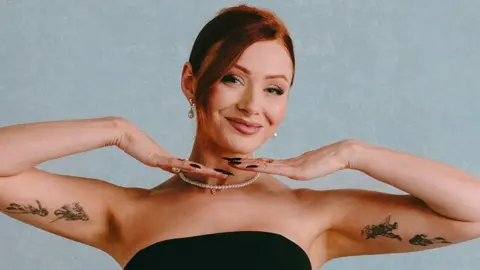 Elz the Witch poses in front of a light blue background. She's wearing a strapless black gown and a thing string of pearls. She's smiling, her hands under her chin, framing her face, and showing off the tattoos of figures drawn in intricate black ink lines on the inside of her upper arms.