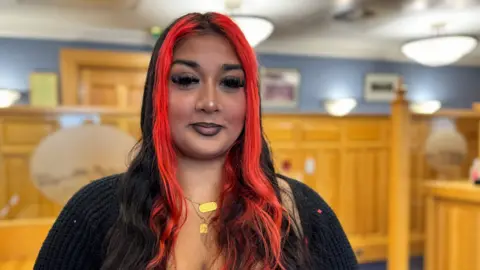 Riona Zaman has long black hair with two bright red streaks at the front framing her face. She is wearing a gold necklace and black cardigan. The room around her is panelled in pine wood with blue at the top part of the wall and several bright white lights.