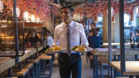 HM Treasury Former Chancellor Rishi Sunak carries two plates of food in a branch of Wagamama's to promote the "Eat Out To Help Out" scheme he launched during Covid.