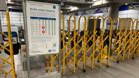 Yellow barriers pulled across ticket barriers with a notice board displaying info under a 'strike action impacts' banner