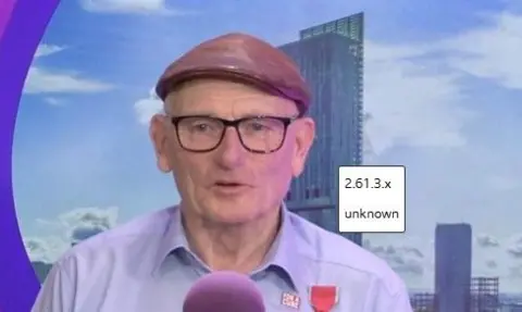 Frank Rothwell is in a radio studio, wearing a flat leather cap and lilac shirt