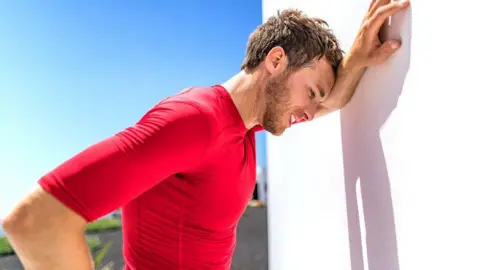 A sweating man with brown hair and wearing a red top leans against a white wall in the sun because he is overheating.